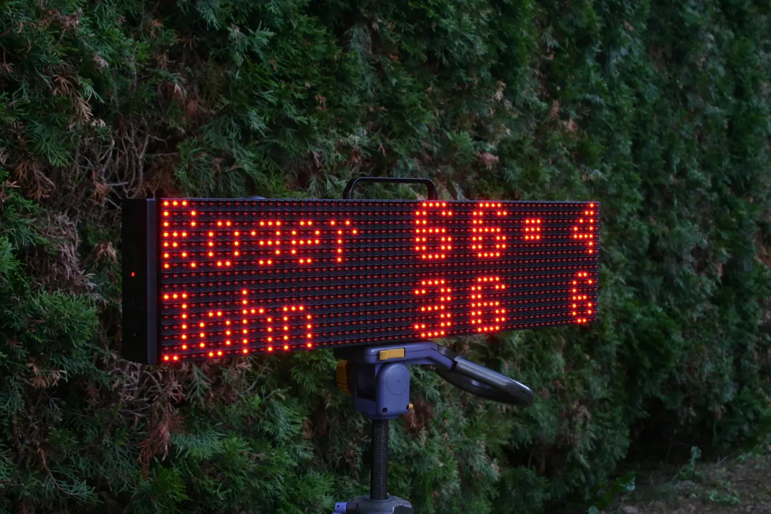 ScreenBattle scoreboard displaying a match score in front of a hedge beside the tennis court